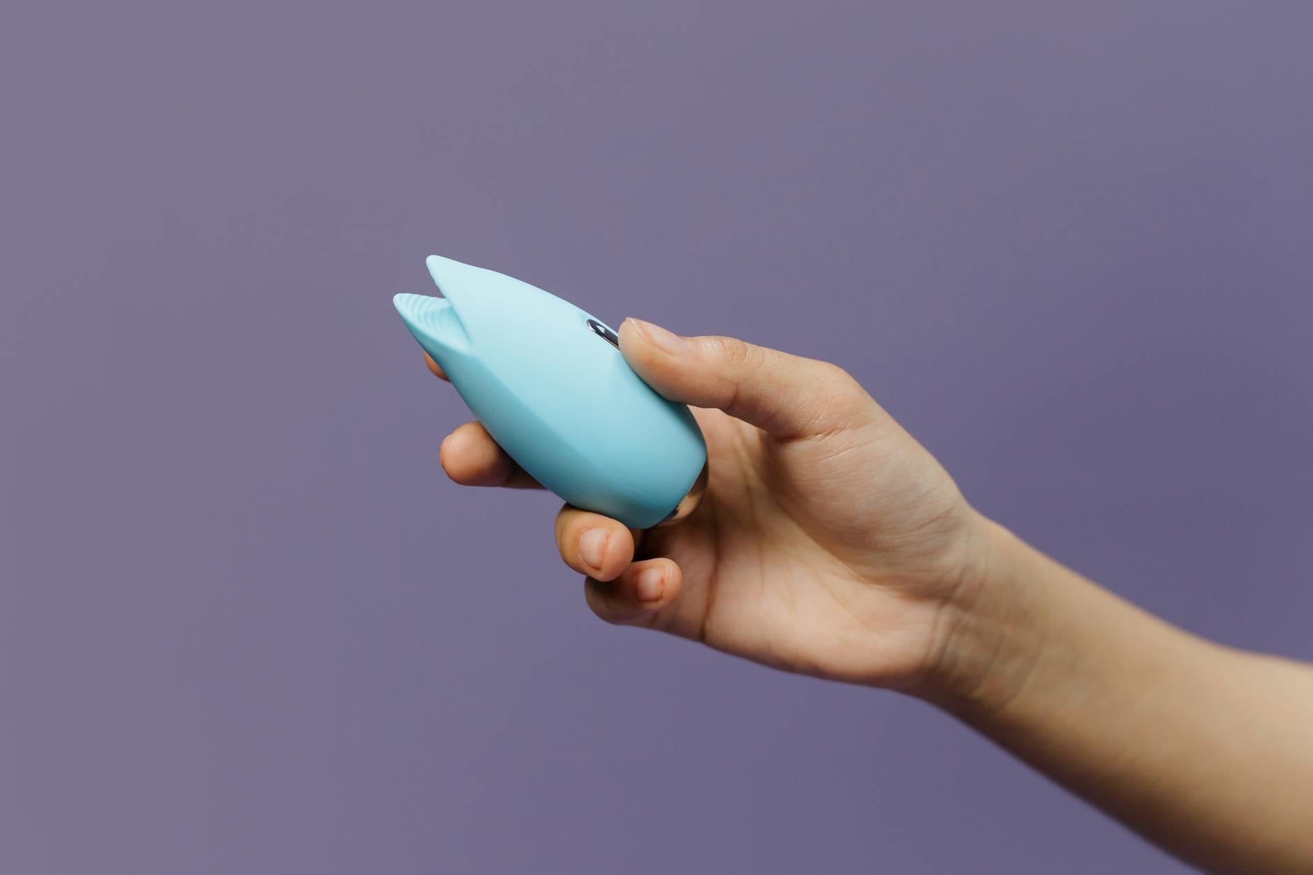 Blue silicone clitoral vibrator in hand against purple background, symbolizing self-pleasure and sexual wellness